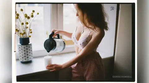 Young woman in white lace lingerie and pink pajama pants drinking water by a bright window in a cozy morning mood.