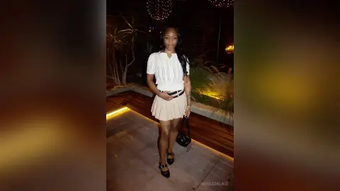 Smiling Black woman in a white lace blouse and beige skirt standing on a patio at night.