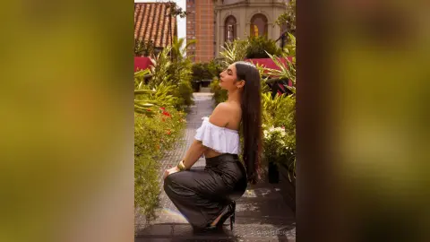 AlmaWhitmore poses outdoors in a white off-the-shoulder crop top and black skirt, looking up with her long dark hair hanging down her back.