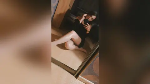 MaragaretSvedine sits on a floor in a black outfit, taking a mirror selfie with her legs stretched out.