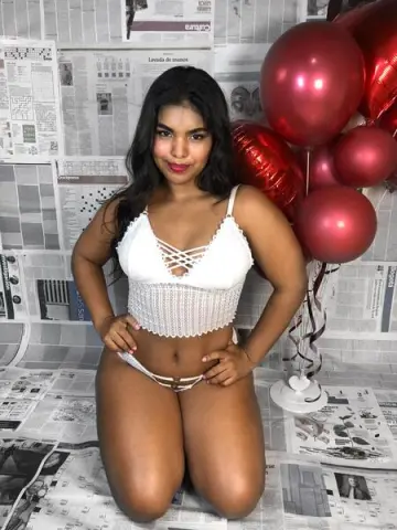 Curvy woman in white lace lingerie posing against a newspaper backdrop with red balloons.