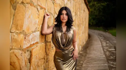 Elegant woman in a metallic gold satin dress posing against a rustic stone wall in a serene outdoor setting.