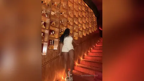 Woman in a white romper standing on a staircase next to a backlit vintage liquor bottle display in a warm lounge.