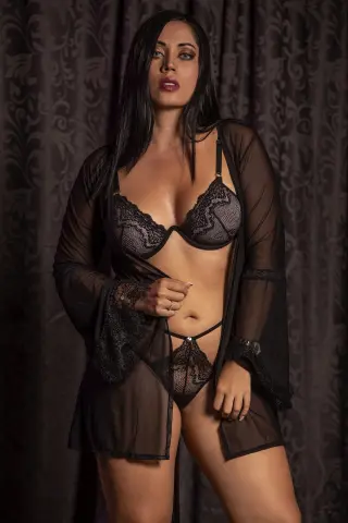 Elegant woman in black lace lingerie and sheer robe posing against a dark patterned backdrop.