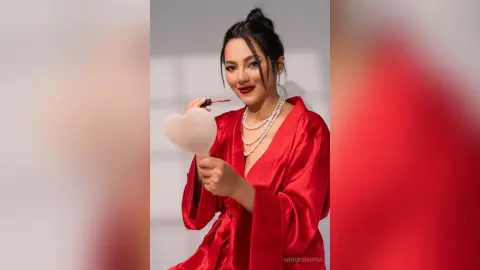 Smiling woman in a red silk robe holding a white heart balloon against a studio background.