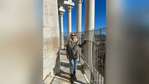 Stylish woman in a grey blazer and jeans standing on a sunny classical stone balcony with tall columns.