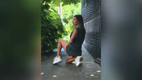Stylish woman in a black tank top and denim shorts kneeling on a city sidewalk near lush green foliage.