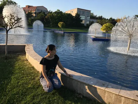 Young woman in a black top and patterned blue pants sitting by a decorative fountain pond in a sunny park.