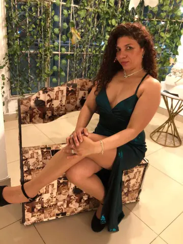 Woman in a glamorous dark green evening dress sitting on a vintage patterned chair in a bright indoor setting.