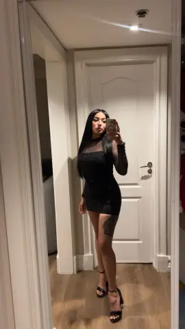 Young woman in a black sparkly mini dress taking a mirror selfie in a modern white hallway.