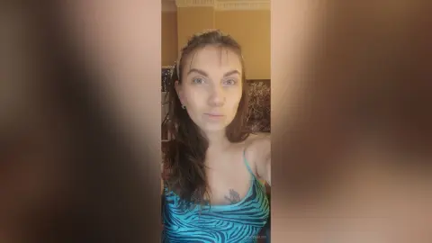 StacyMalvin wearing a blue patterned tank top, looking directly at the camera with her long brown hair down.