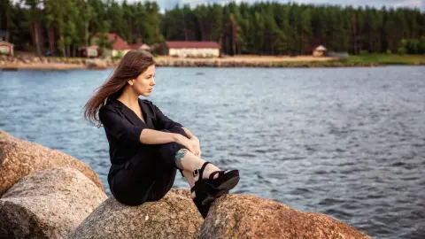 Young woman in black outfit sitting on rocks by a lake near a forest during a peaceful summer day.