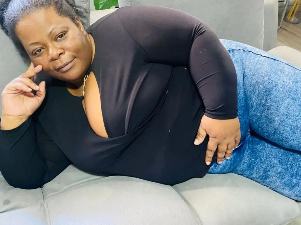 Smiling Black woman in a black long-sleeve top and blue jeans relaxing on a grey sofa.