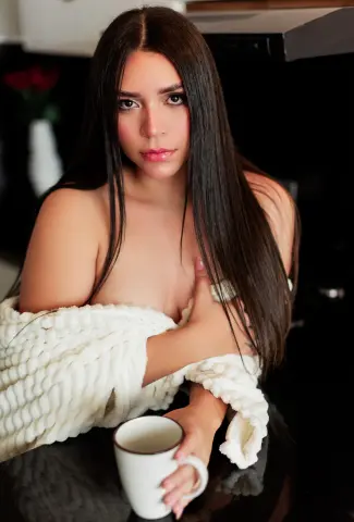 Young woman with long dark hair wearing a cozy white knit sweater holding a mug in a moody kitchen setting.