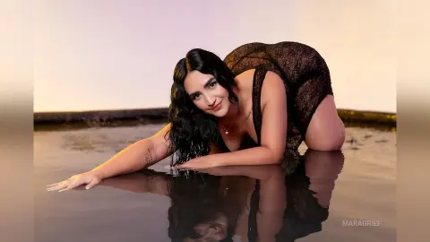 MaraGrief poses in a black mesh bodysuit while kneeling in shallow water, looking at the camera with long dark hair.