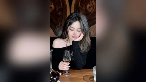 Melancholic young woman in a black off-the-shoulder top holding a glass of white wine at a dimly lit bar.