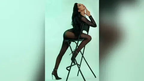 NicoleBella sits on a black chair wearing black sheer lingerie and tights, posing with her head tilted back against a light green background.