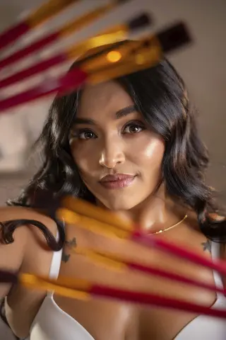Smiling woman with wavy dark hair in a white tank top framed by blurred makeup brushes in a soft studio setting.
