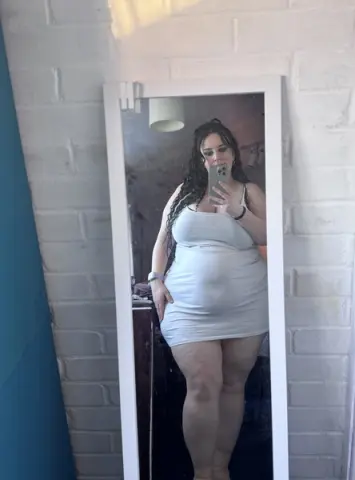 Curvy woman in a white mini dress taking a mirror selfie in a bright, minimalist room.