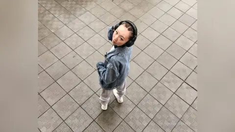 Young woman in denim jacket and headphones standing on a tiled floor looking up with a calm expression.