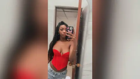 NatyGlow poses in a red corset and light denim jeans, showing off her long dark hair in a mirror selfie.