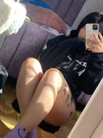 Young woman in a black Tommy Hilfiger hoodie sitting on a purple sofa for a cozy indoor mirror selfie.