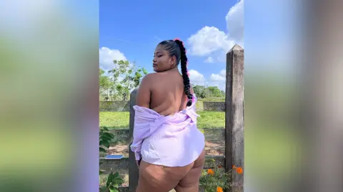 Curvy woman in a light pink shirt posing outdoors against a sunny sky and green field.