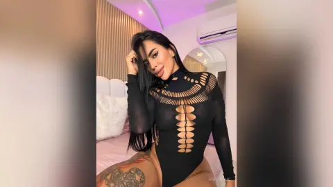 Glamorous woman in a black long-sleeve cutout bodysuit posing in a modern bedroom with soft lighting.