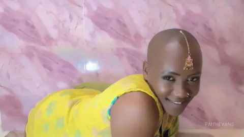 Smiling bald woman in a bright yellow patterned dress posing against a pink marble wall background.