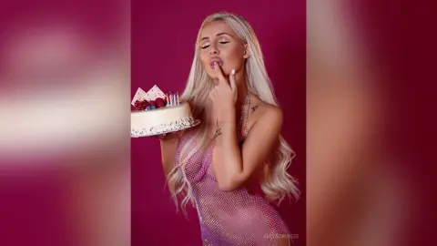 Anita licks her finger while holding a white frosted cake, wearing a sheer pink mesh bodysuit against a deep red background.