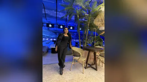 Stylish woman in a black outfit standing in a modern lounge with tropical plants and blue ambient lighting.