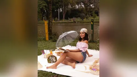 Young woman in a pink off-the-shoulder top relaxing on a picnic blanket by a lake with an umbrella.