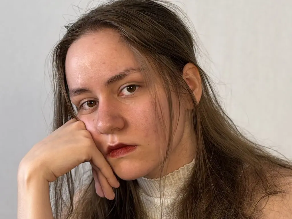 Young woman with long brown hair wearing a white turtleneck sweater in a minimalist indoor setting.