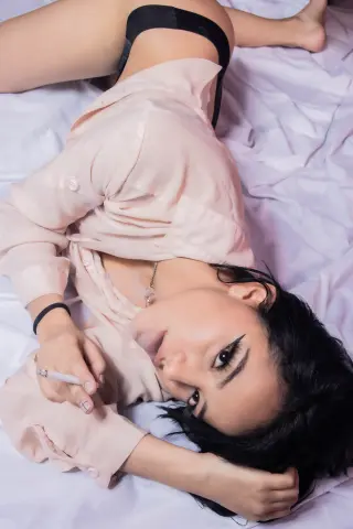 Young woman in a beige silk shirt lying on white bedsheets with a relaxed, moody aesthetic.