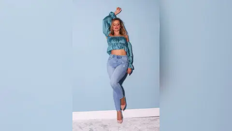 Gia poses against a blue wall wearing a teal silk long-sleeve crop top and light blue denim jeans.
