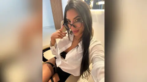 Smiling woman in glasses and white button-down shirt relaxing in a bright, modern indoor setting.