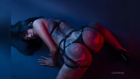 Tattooed woman in black strappy lingerie posing on a dark blue floor with moody red lighting.