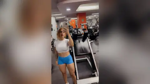 Blonde woman in white crop top and blue shorts taking a mirror selfie in a modern gym setting.