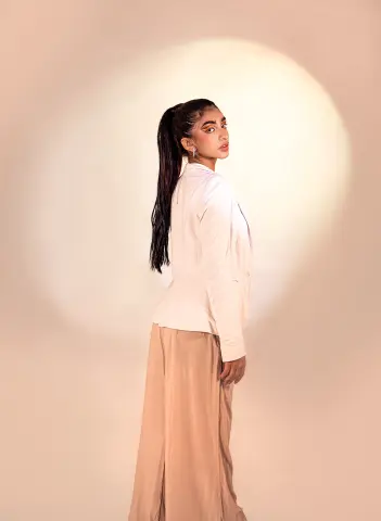 Veronica_Hottiee poses sideways in a white blazer and tan wide-leg trousers against a warm, lit background.