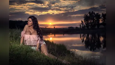 MissyJolie sits in a grassy field by a lake at sunset, wearing a light pink off-the-shoulder top and white shorts.