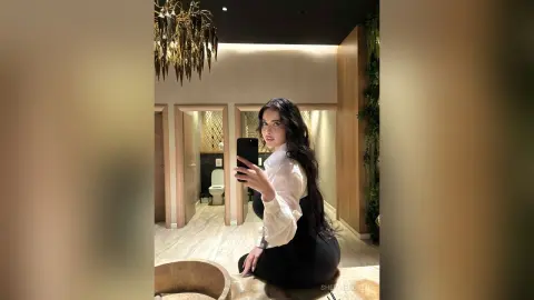Elegant woman in a white blouse taking a mirror selfie in a modern, luxury bathroom with gold decor.