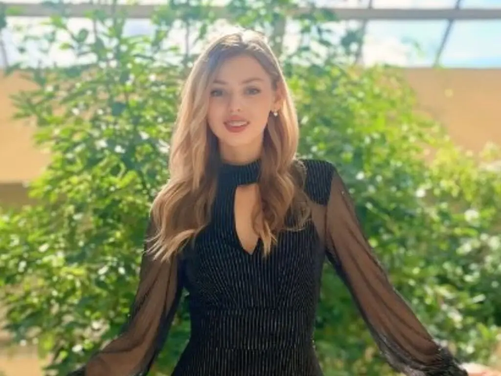 Blonde woman in a black long-sleeve cutout dress smiling in a bright sunlit garden setting.