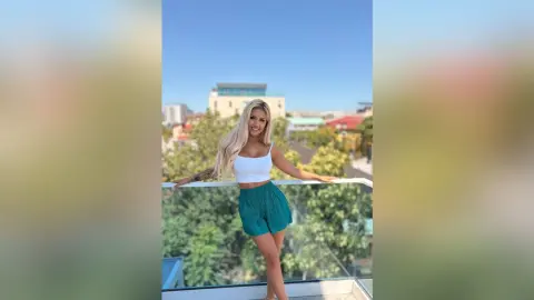 Anita stands on a balcony wearing a white tank top and green shorts, looking at the camera with long blonde hair.