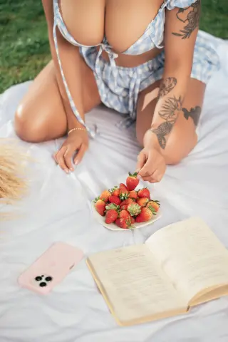 Woman in blue gingham bikini relaxing on a white blanket with strawberries and an open book outdoors.