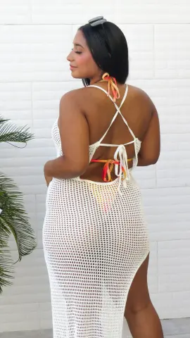 Woman in a white crochet maxi beach cover-up standing against a white tiled wall in bright sunlight.