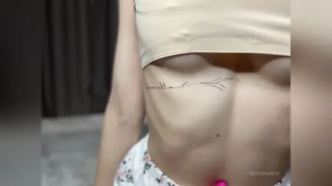 Close-up of a woman in a beige crop top showing a delicate script tattoo on her ribcage in a soft indoor setting.