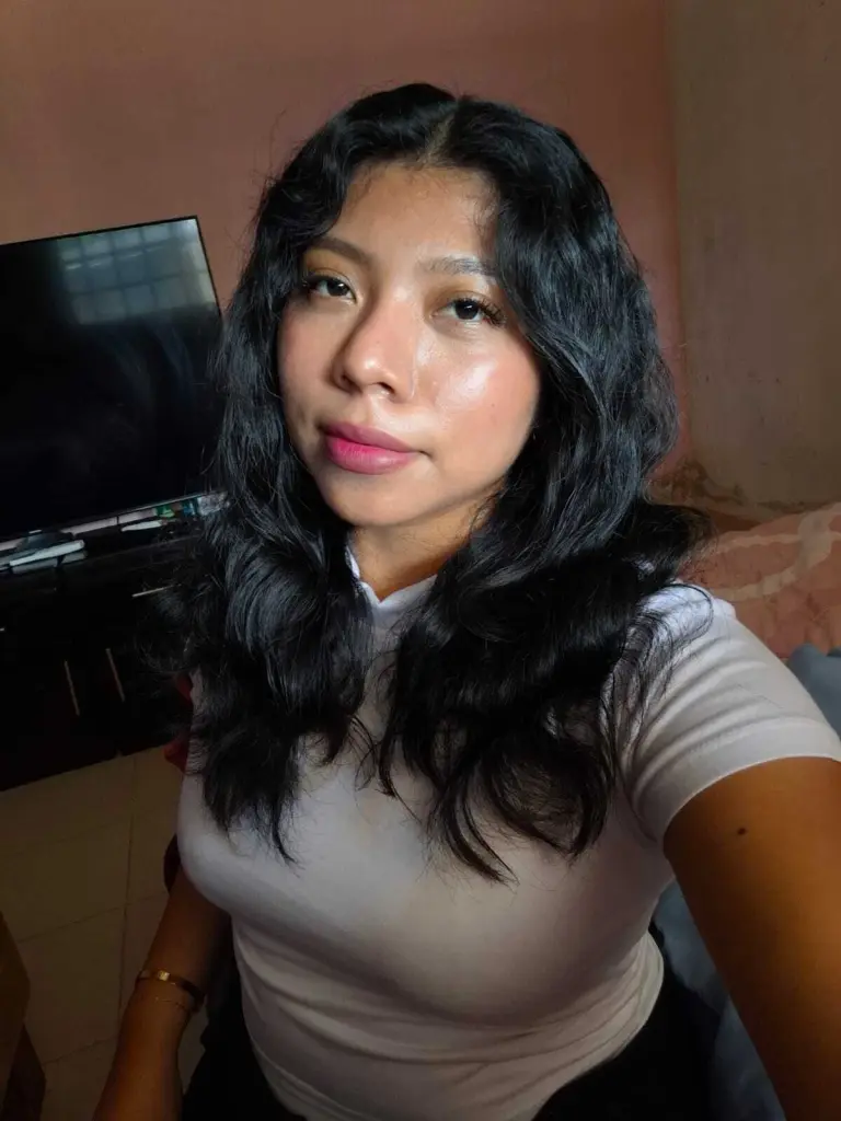 Yoyo_Noor poses with long dark hair, wearing a purple off-the-shoulder top against a light curtain background.