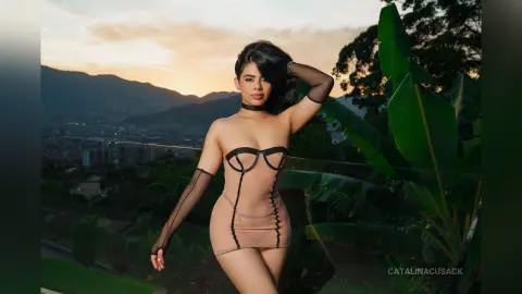 Glamorous woman in a beige corset mini dress posing against a tropical sunset mountain landscape.