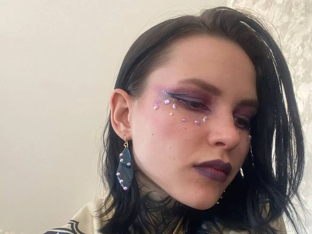 AndreaHazel looks down with dark purple lipstick and glittery eye makeup, wearing large blue stone earrings and a patterned top.