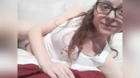 Smiling woman in a white tank top relaxing on a white bed in a cozy indoor setting.